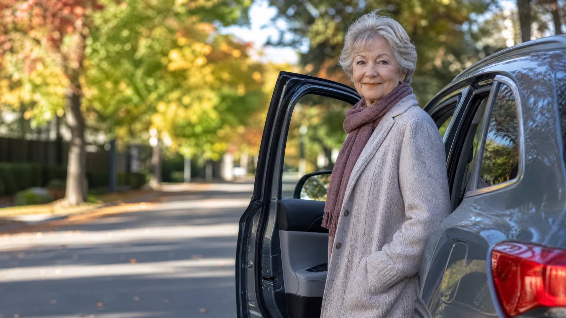 An older woman in a light coat and pink scarf stands beside an open rear door of a dark car on a tree-lined street with autumn foliage.