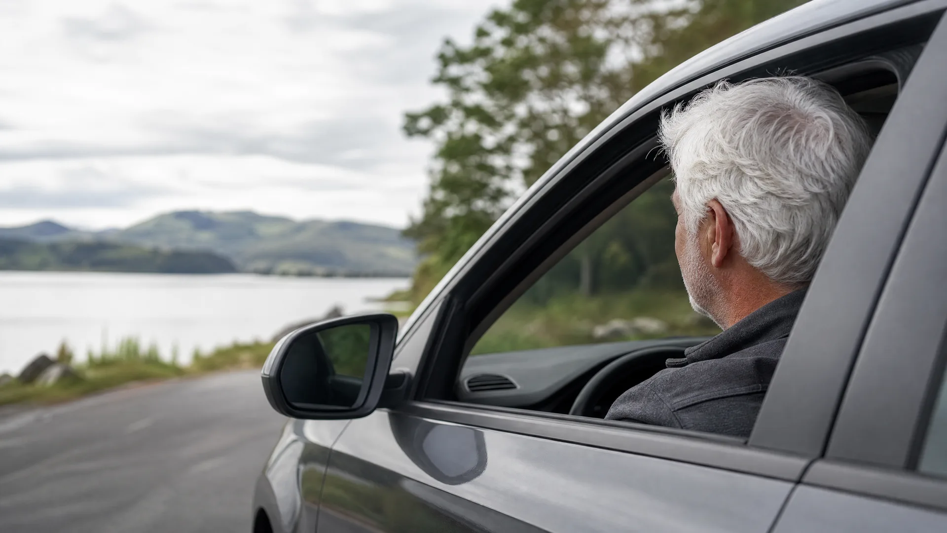 A white-haired man sits in the driver's seat of a grey car with the window down, parked on a road beside a lake with hills in the background under an overcast sky.