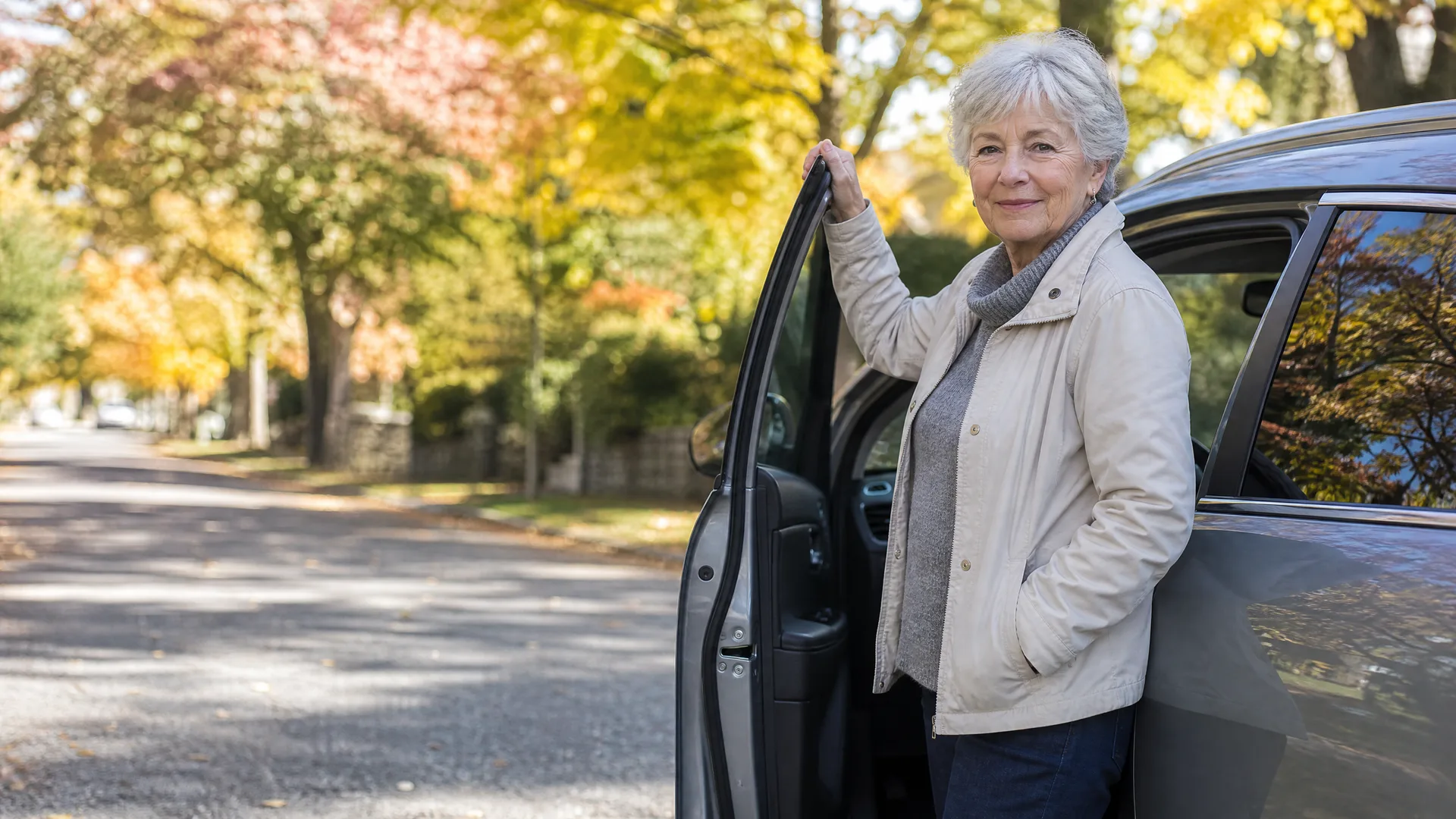 An older woman with short white hair in a beige jacket stands holding the open front door of a dark car parked on a tree-lined street with yellow autumn foliage.